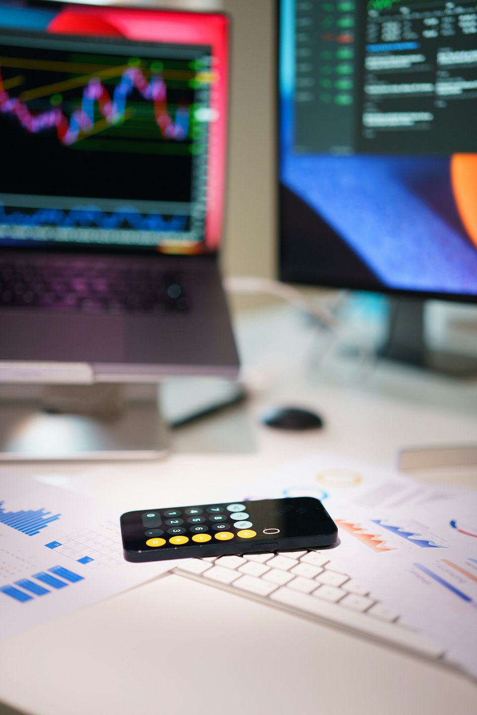 Smartphone auf Finanzdokumenten neben Laptop mit Börsencharts.